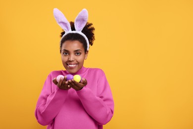 Happy African American woman in bunny ears headband holding Easter eggs on orange background, space for text