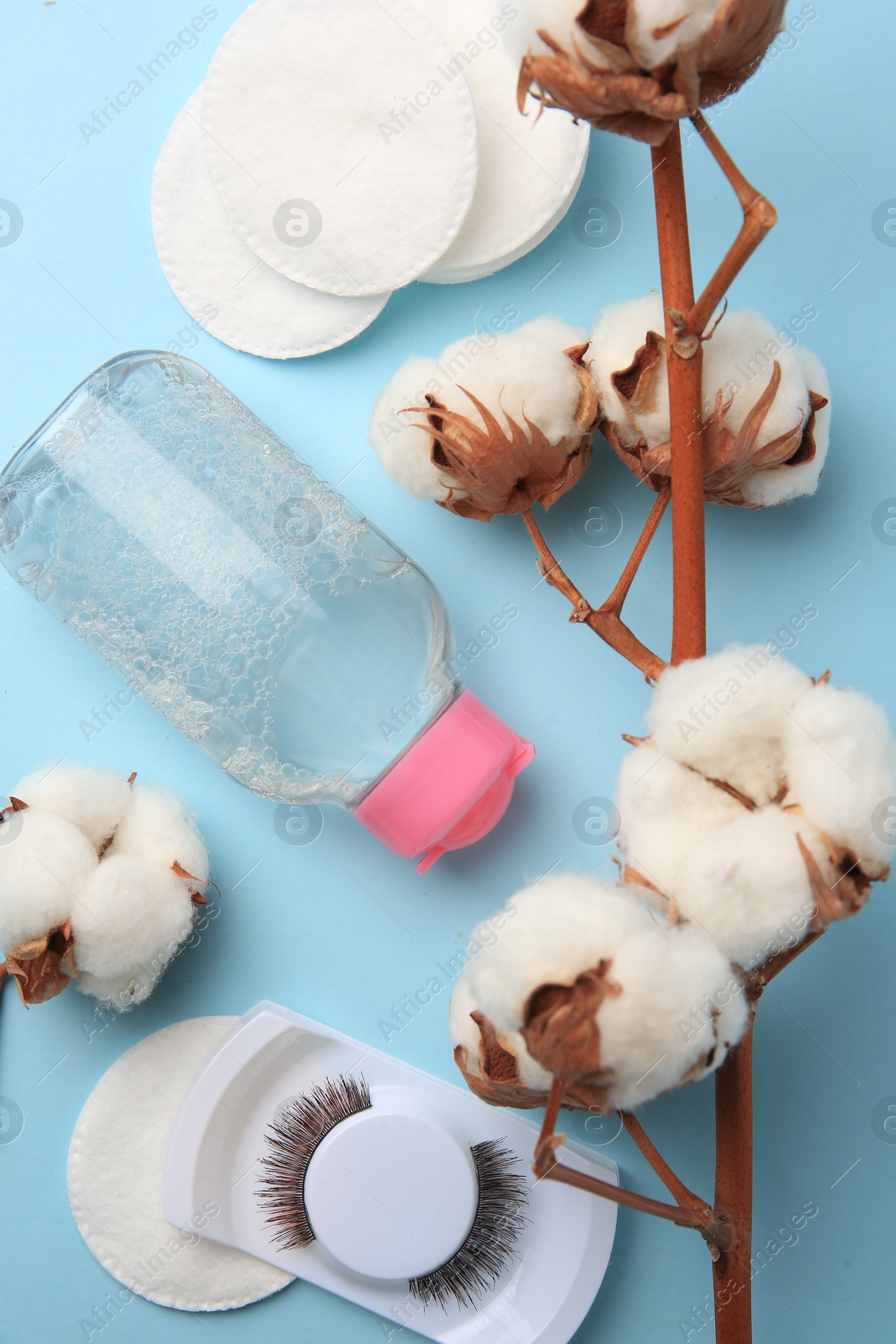 Photo of Bottle of makeup remover, cotton flowers, pads and false eyelashes on light blue background, flat lay