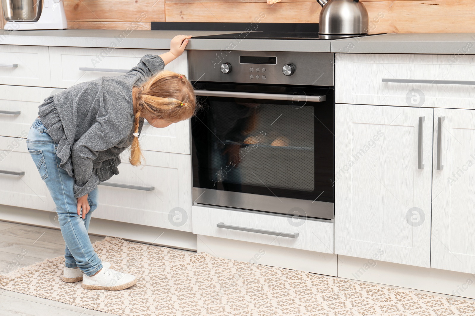 Photo of Little girl waiting for preparation of cookies in oven at home