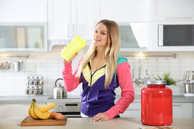 Photo of Young woman drinking protein shake near table with ingredients in kitchen