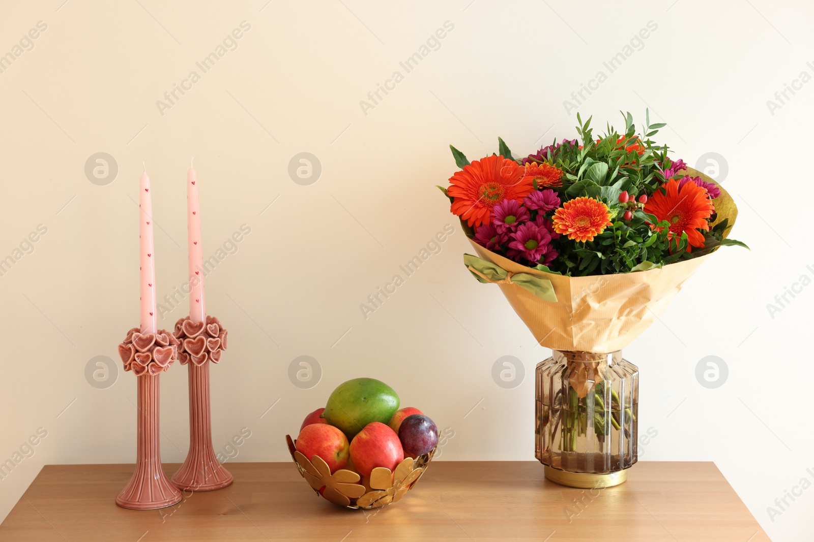 Photo of Bouquet of flowers, bowl with fresh fruits and candles on wooden table near white wall