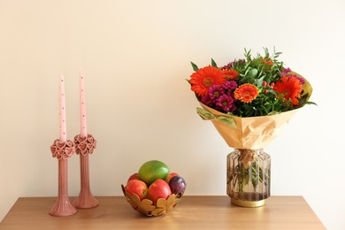 Photo of Bouquet of flowers, bowl with fresh fruits and candles on wooden table near white wall