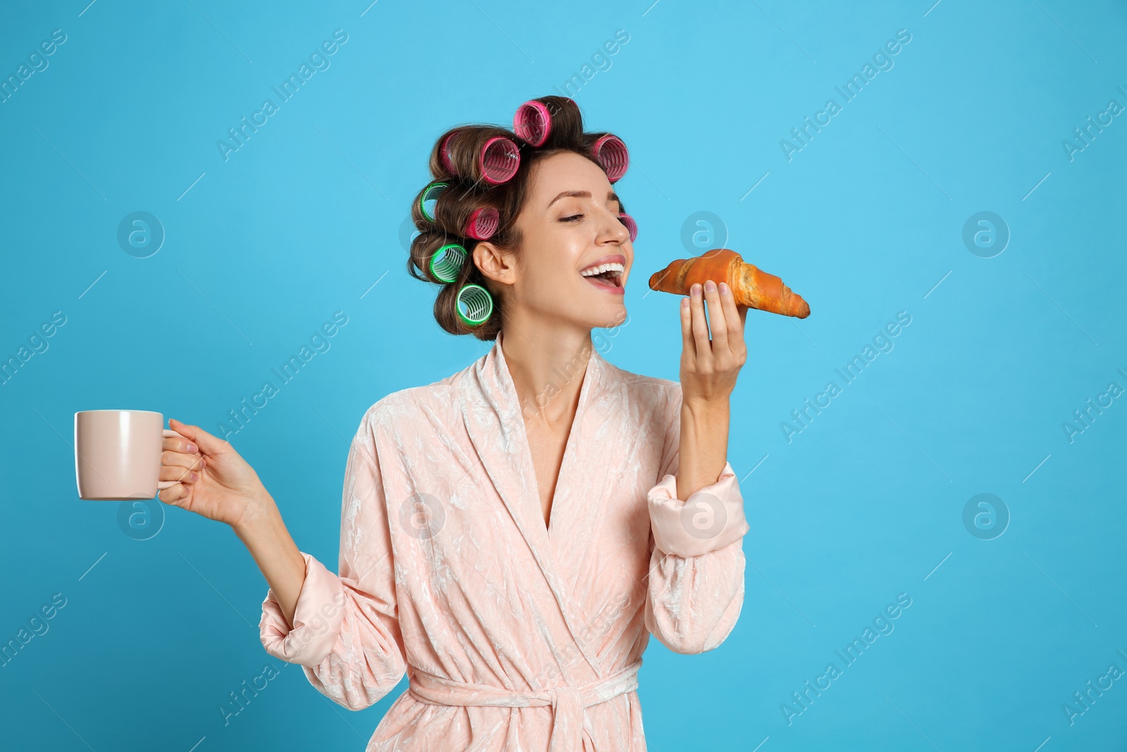 Photo of Happy young woman in bathrobe with hair curlers holding croissant and cup of drink on light blue background