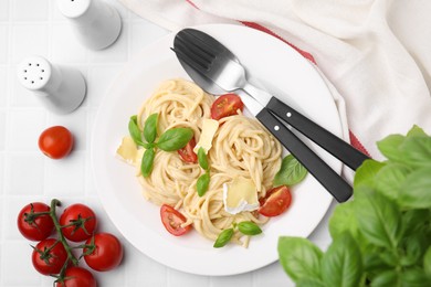 Delicious pasta with brie cheese, tomatoes, basil and cutlery on white tiled table, flat lay