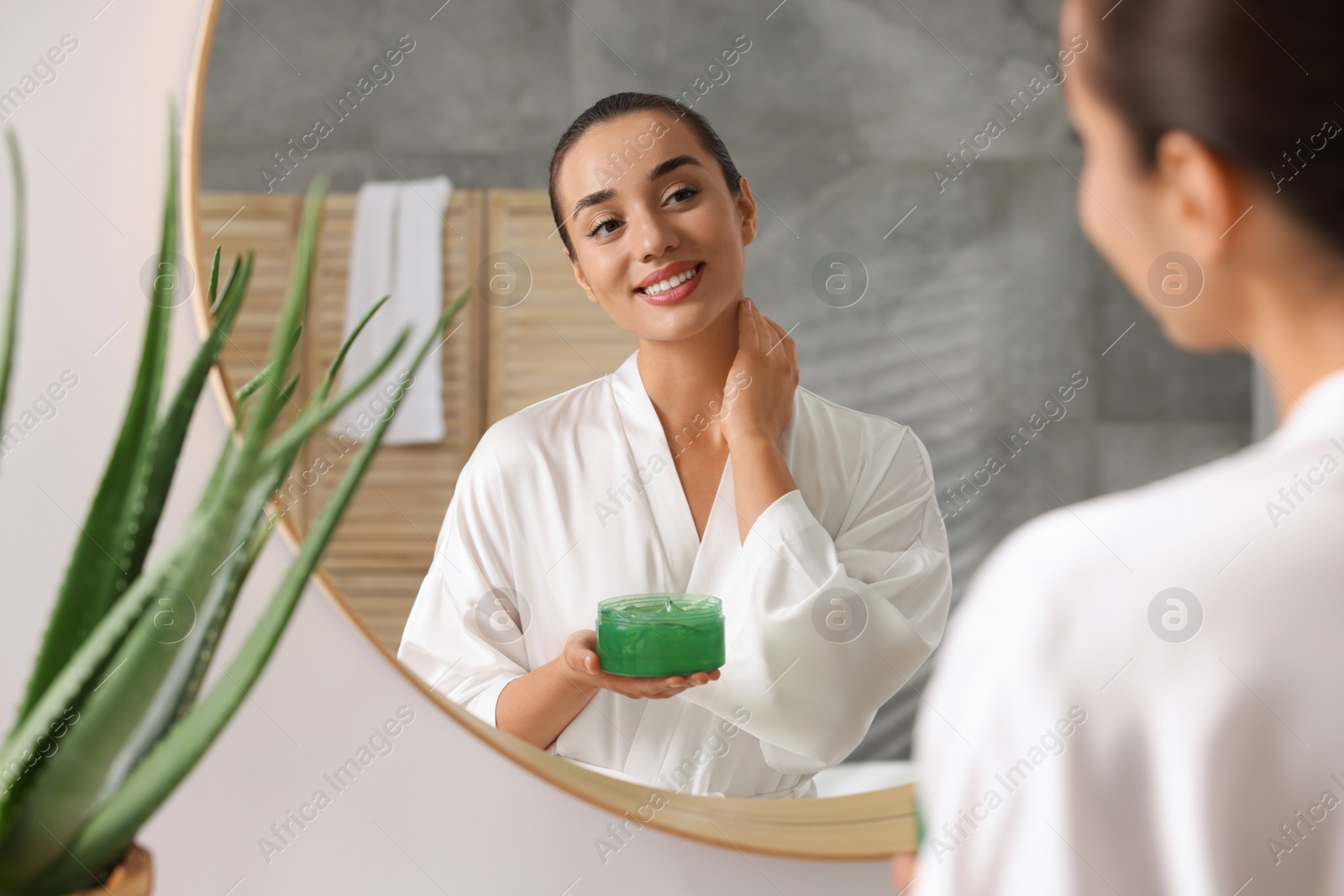 Photo of Young woman holding jar of aloe gel near mirror in bathroom