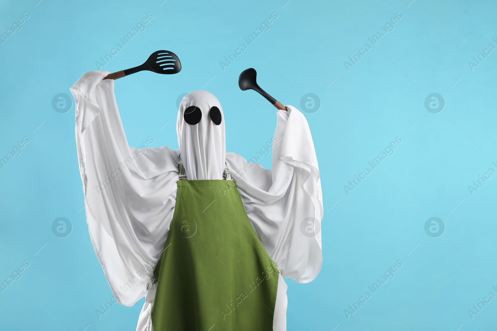 Photo of Woman in ghost costume and apron with kitchen set on light blue background, space for text
