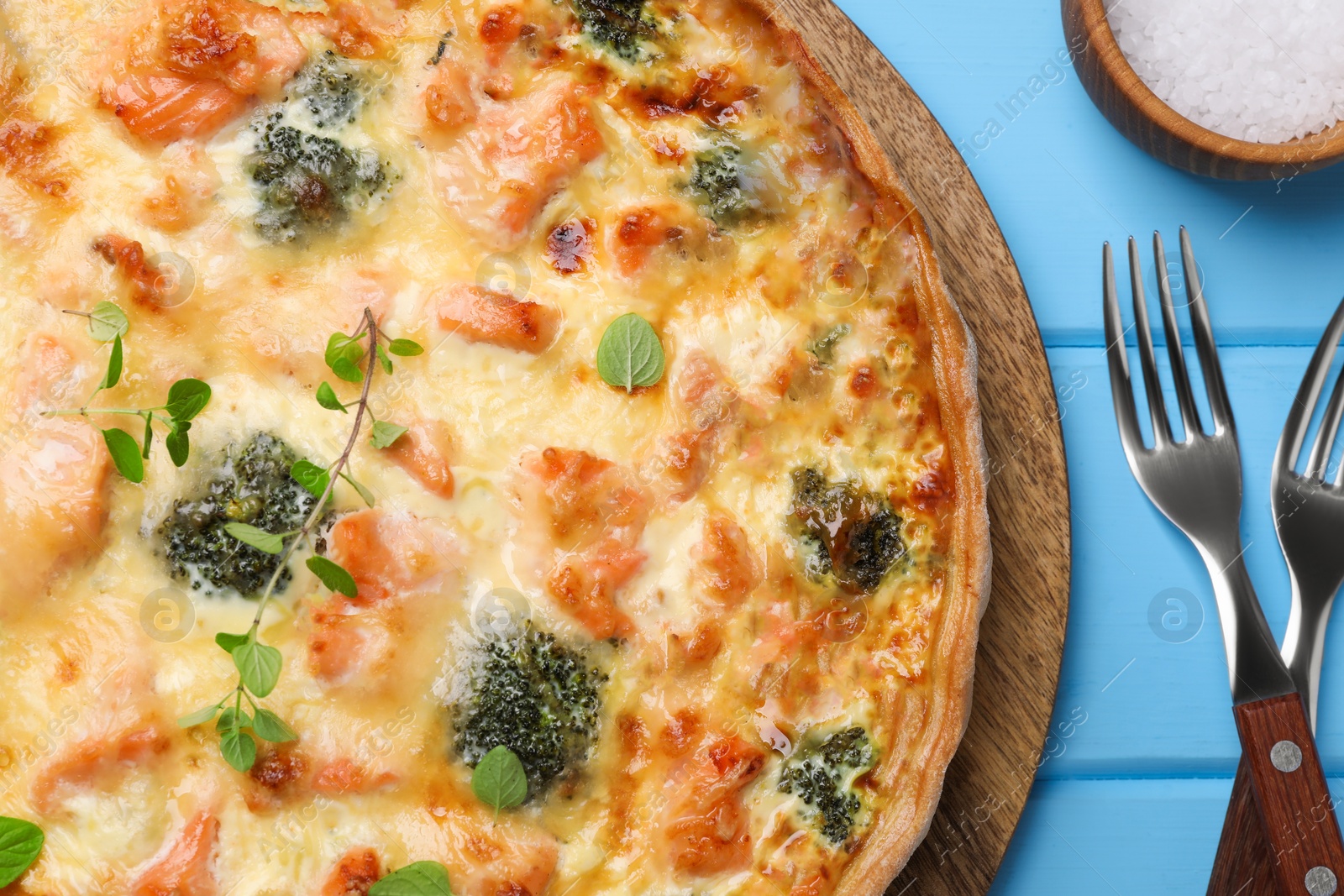 Photo of Delicious homemade salmon quiche with broccoli, forks and salt on light blue wooden table, flat lay