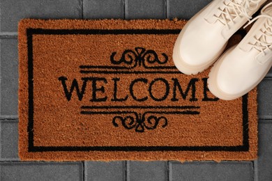 Door mat with word Welcome and stylish boots on floor, flat lay