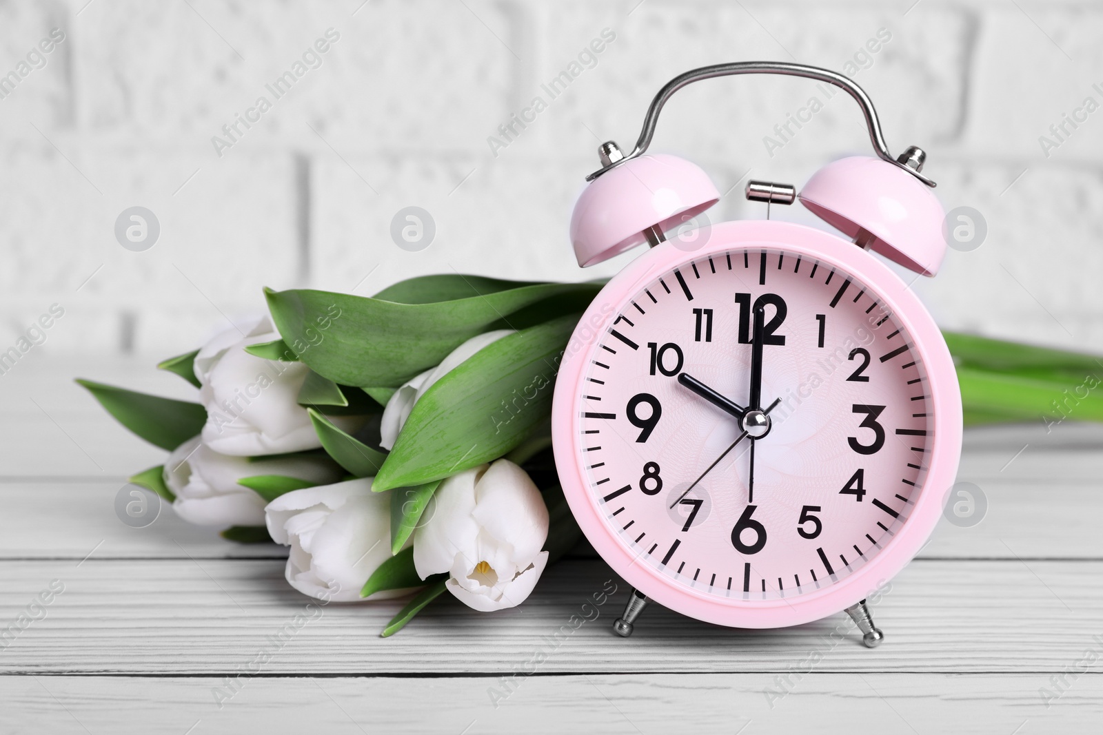 Photo of Pink alarm clock and beautiful tulips on white wooden table against brick wall, closeup. Spring time