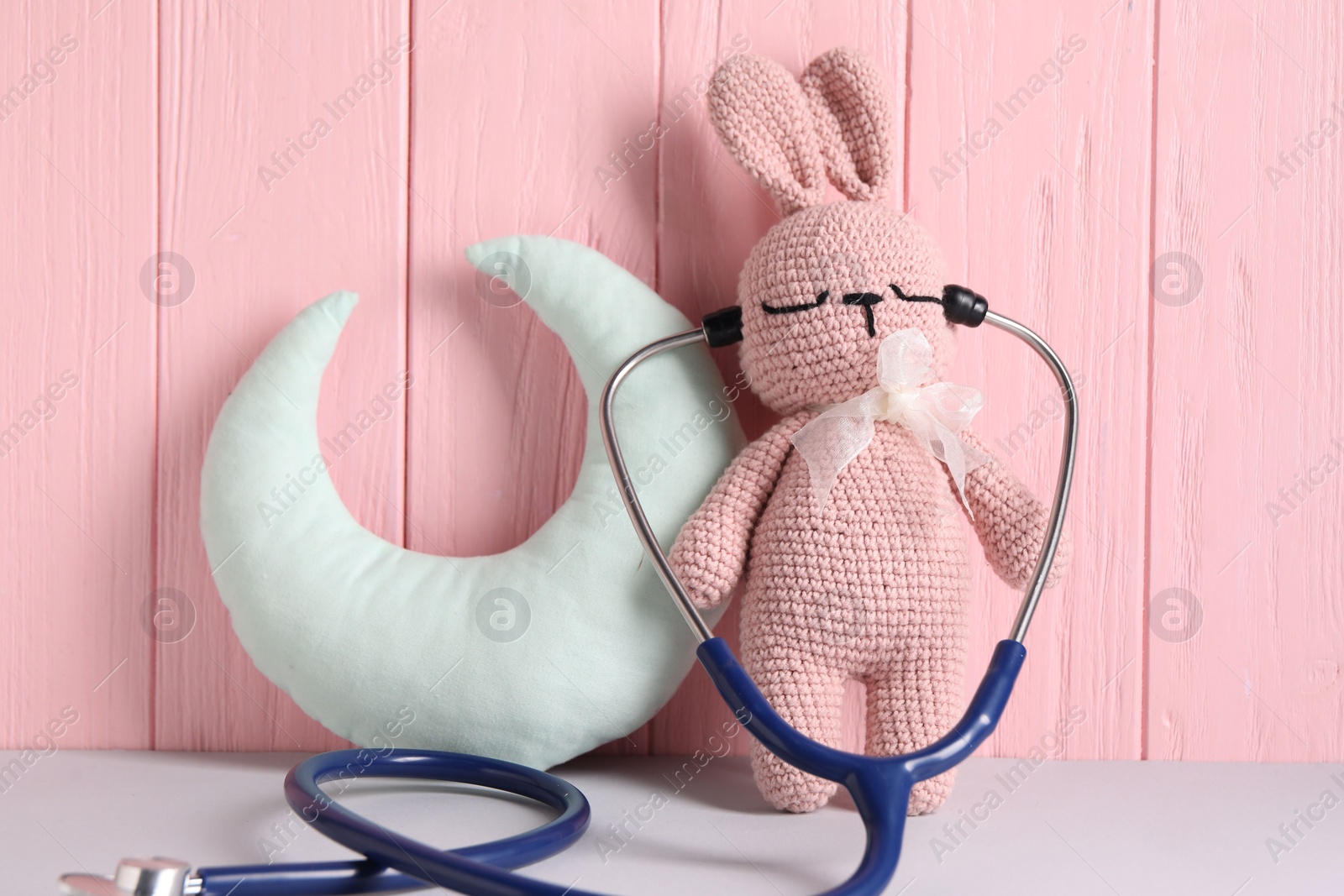 Photo of Toy bunny with stethoscope and pillow on light table near pink wooden wall