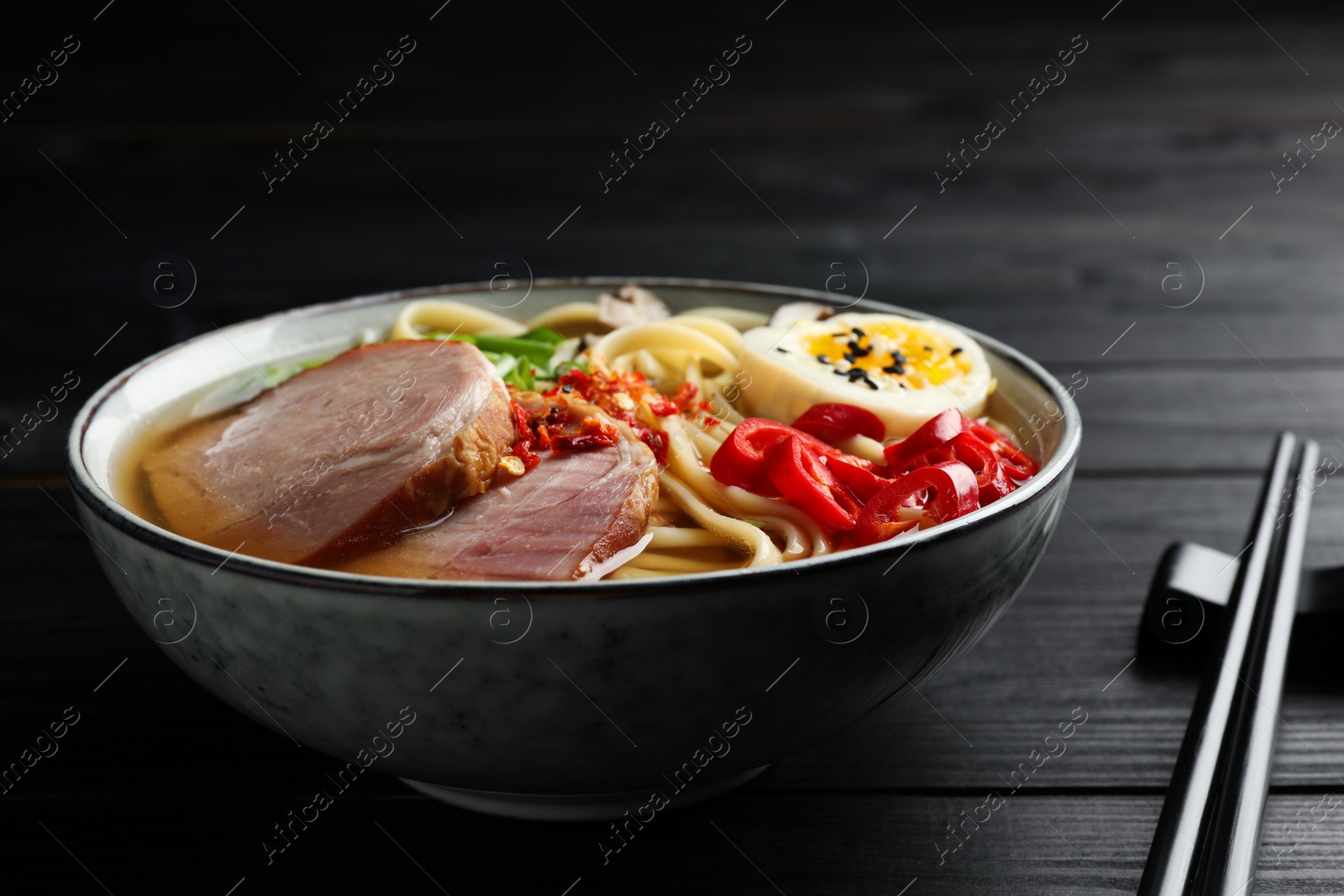 Photo of Delicious ramen in bowl and chopsticks on black wooden table, closeup. Noodle soup