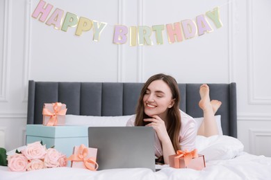 Photo of Beautiful young woman with laptop near gift boxes and rose flowers on bed in room. Happy Birthday