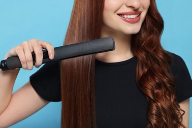 Young woman using hair iron on light blue background, closeup