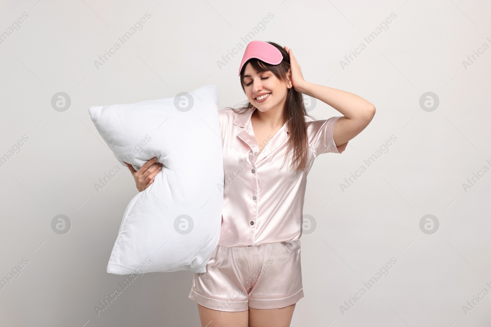 Photo of Happy woman in pyjama and sleep mask holding pillow on light grey background