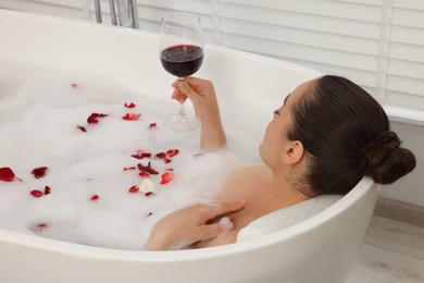 Woman with glass of wine taking bath in tub with foam and rose petals indoors