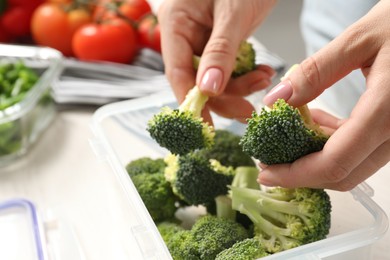 Photo of Woman putting broccoli into plastic container at white table, closeup. Food storage