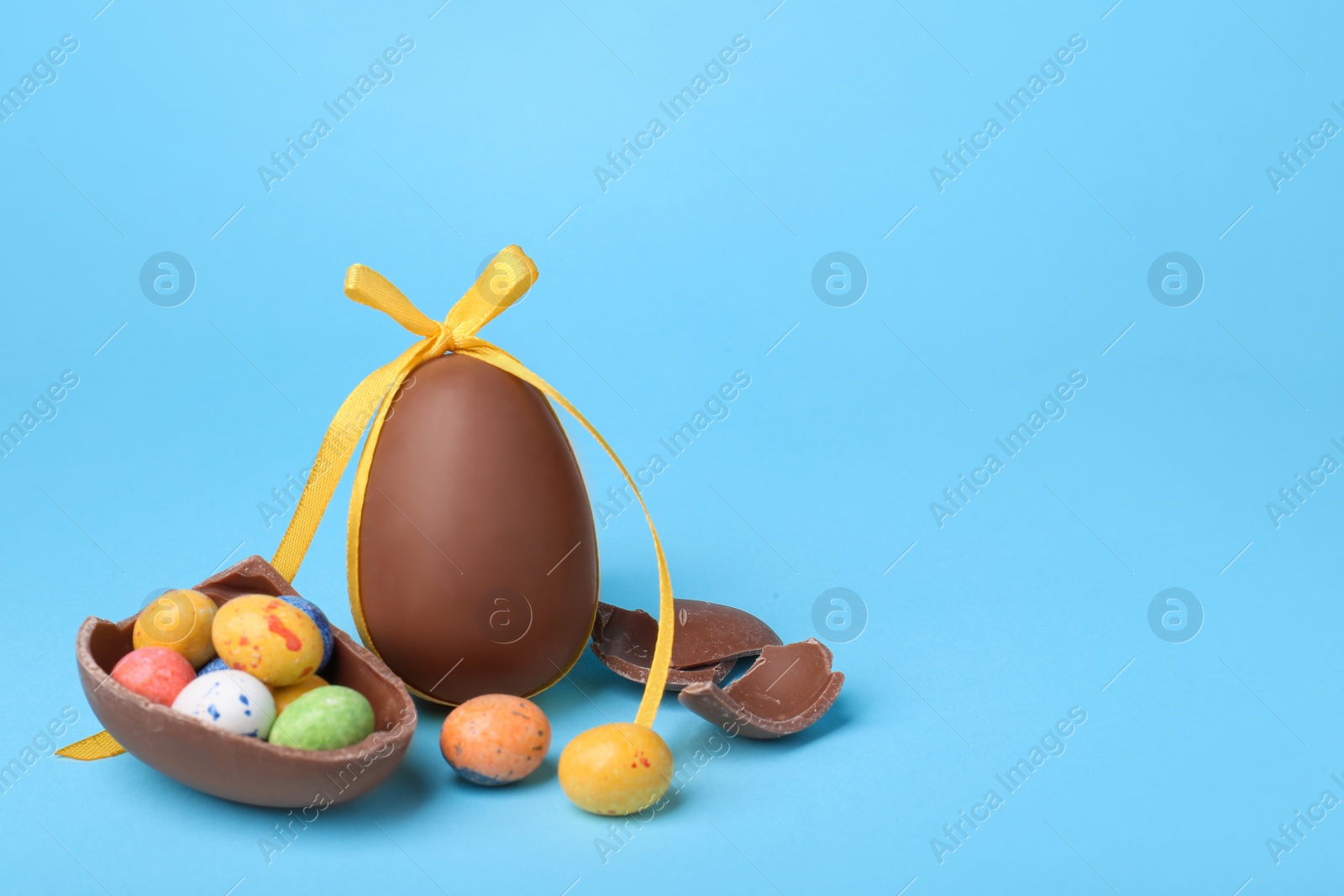 Photo of Tasty whole chocolate egg with yellow bow and different candies on light blue background. Space for text