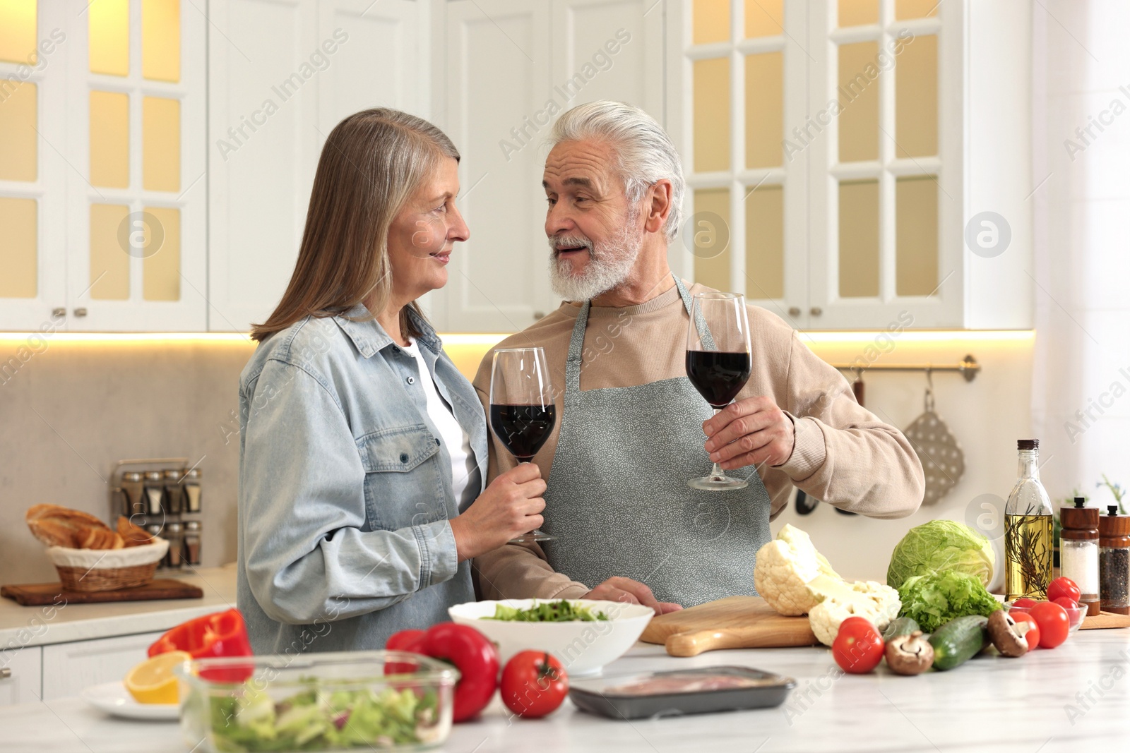 Photo of Happy senior couple with glasses of wine cooking together in kitchen