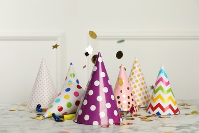 Photo of Shiny confetti falling on marble table with party hats and festive items near white wall indoors