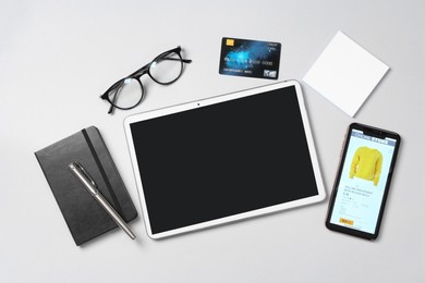 Online store website on device screen. Tablet, smartphone, stationery, glasses and credit card on light grey background, flat lay