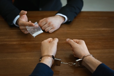 Photo of Police officer interrogating drug dealer in handcuffs at desk indoors. Criminal law