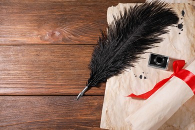 Black feather pen, inkwell and vintage parchment with ink stains on wooden table, flat lay. Space for text