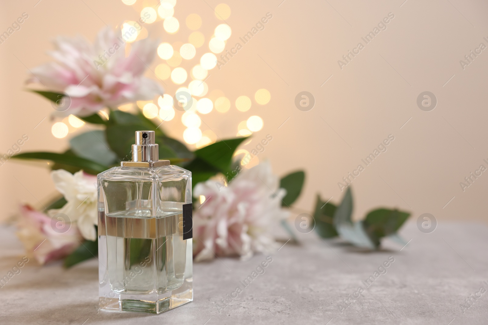 Photo of Bottle of perfume and beautiful lily flowers on table against beige background with blurred lights, space for text