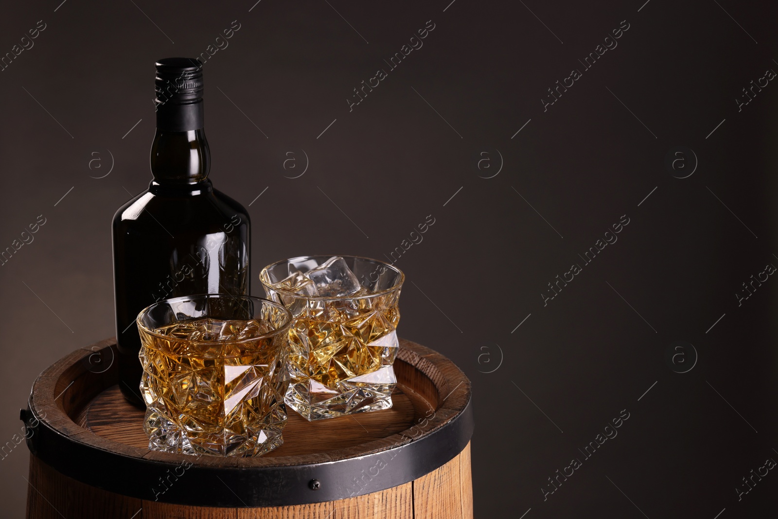 Photo of Whiskey with ice cubes in glasses and bottle on wooden barrel against dark background, space for text