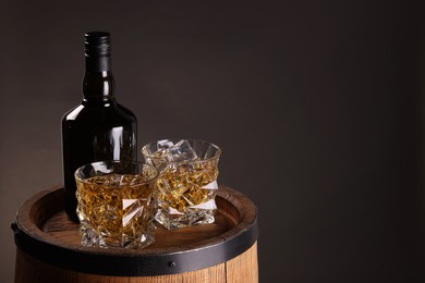 Whiskey with ice cubes in glasses and bottle on wooden barrel against dark background, space for text