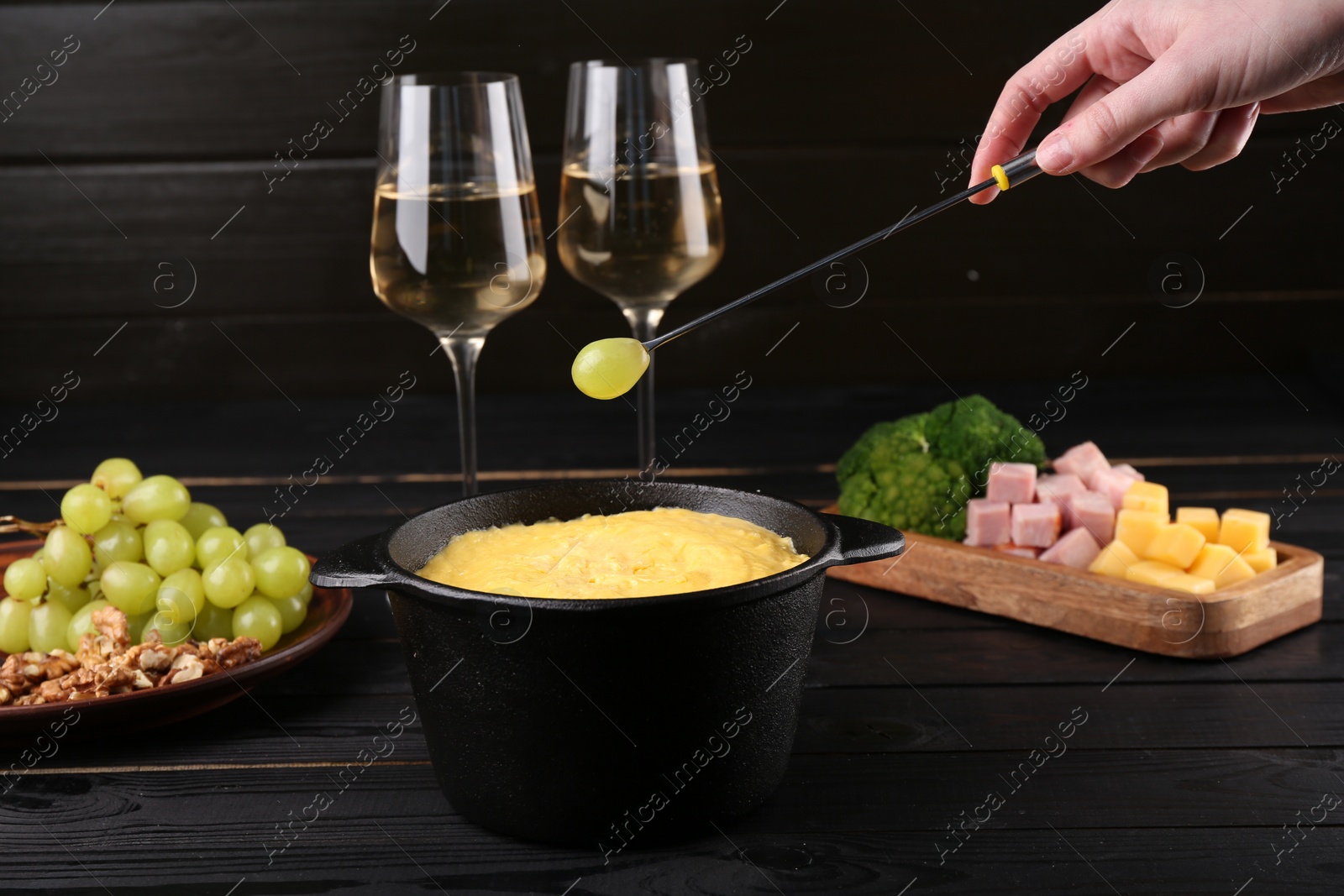 Photo of Woman dipping grape into fondue pot with melted cheese at black wooden table, closeup