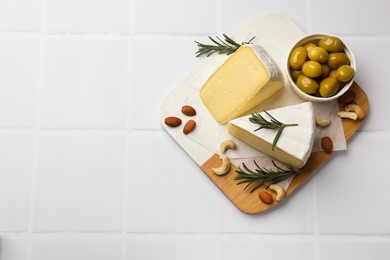 Photo of Pieces of tasty camembert cheese, rosemary, nuts and olives on white tiled table, top view. Space for text