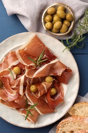 Slices of tasty cured ham, rosemary, bread and olives on blue wooden table, flat lay