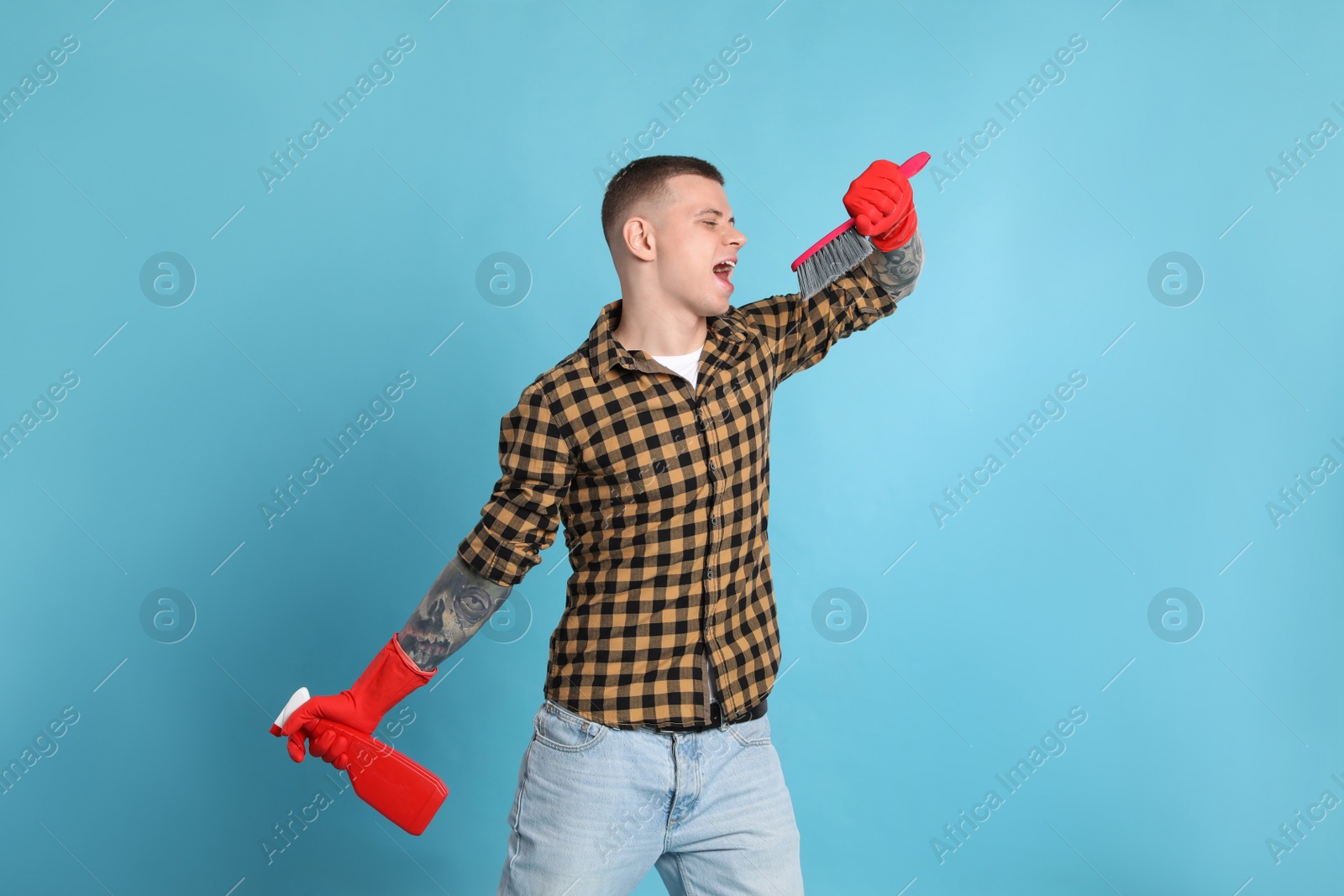 Photo of Handsome young man with brush and bottle of detergent singing on light blue background