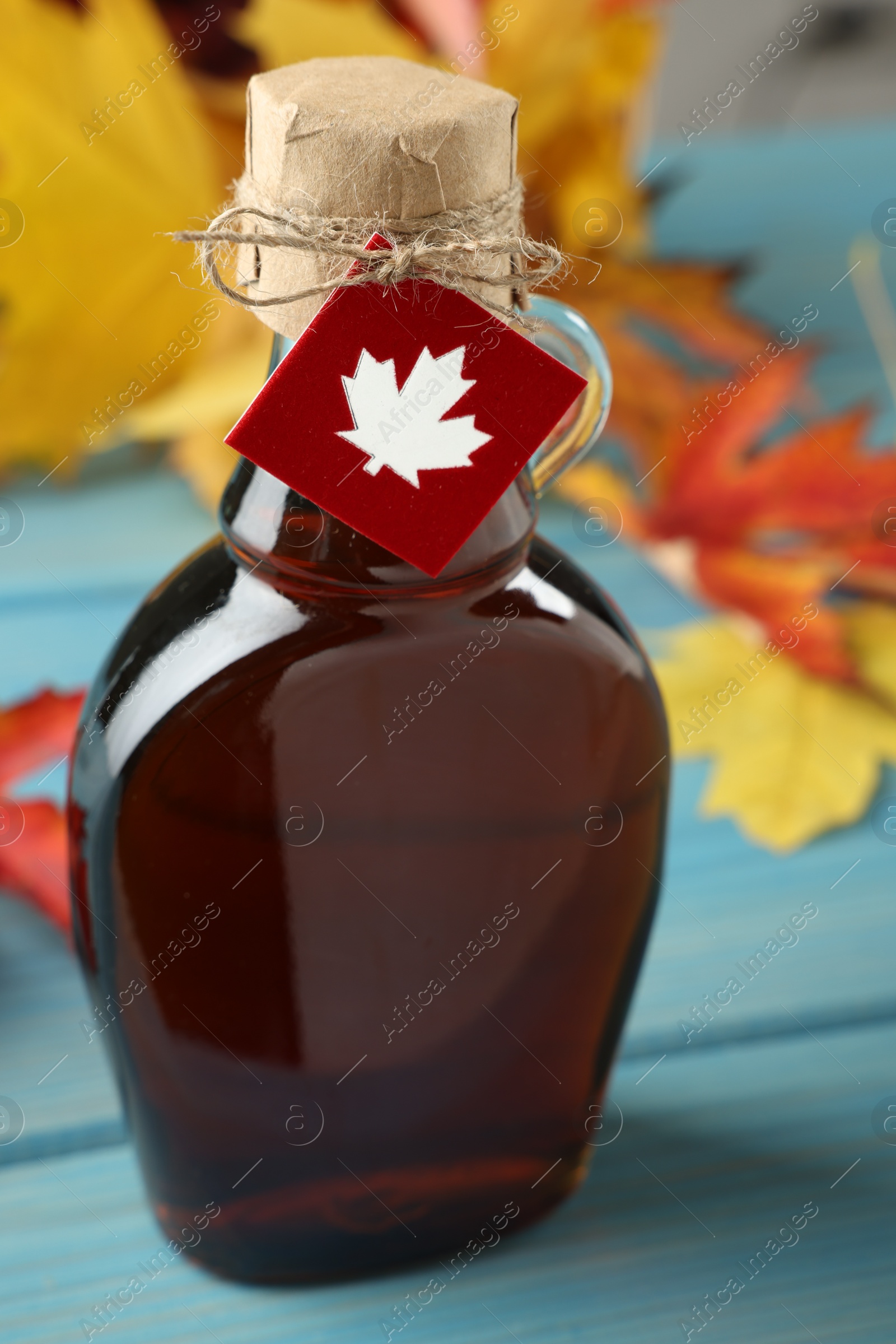 Photo of Glass bottle of tasty maple syrup and dry leaves on light blue wooden table, closeup