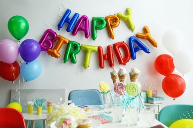 Photo of Table with treats and phrase HAPPY BIRTHDAY made of colorful balloon letters in living room