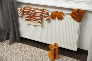 Photo of Heating radiator with knitted hat, sweater, gloves and rubber boots indoors