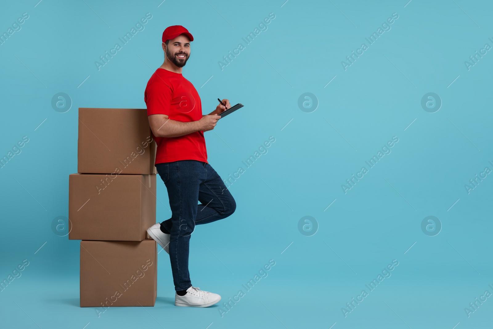 Photo of Happy young courier with clipboard and stack of parcels on light blue background, space for text