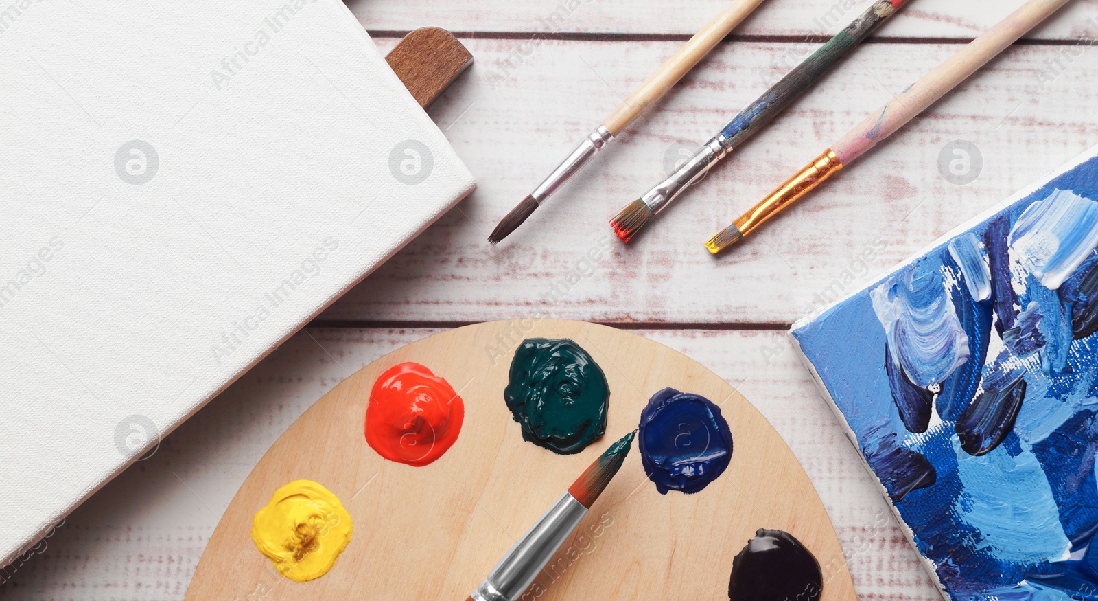 Photo of Artist's palette with samples of colorful paints, brushes and canvases on white wooden table, flat lay