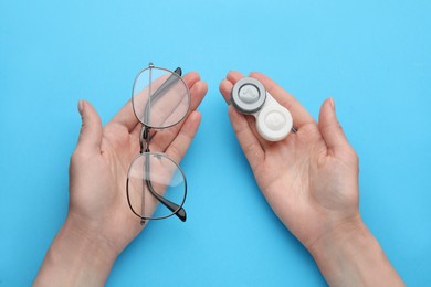 Woman holding case with contact lenses and glasses on light blue background, top view