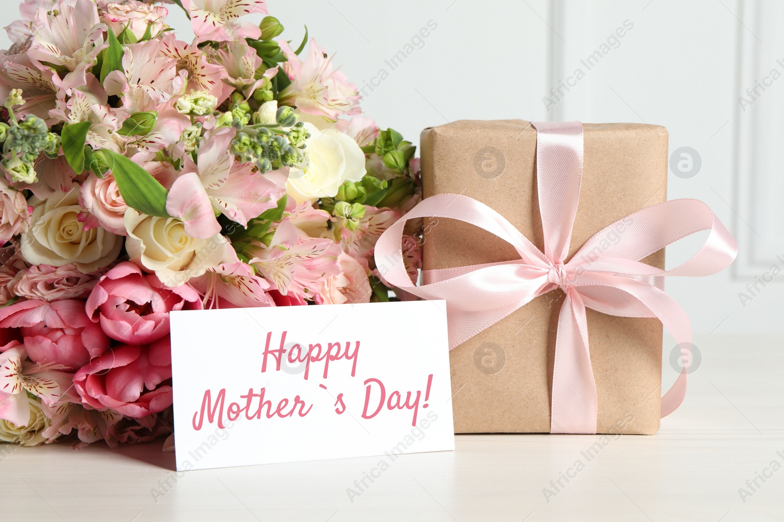 Image of Happy Mother's Day greeting card, gift box and bouquet of beautiful flowers on white wooden table