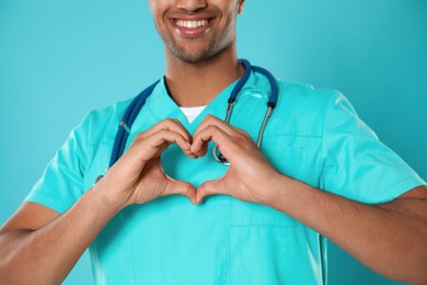 Photo of Doctor or medical assistant (male nurse) in uniform making heart with hands turquoise background, closeup