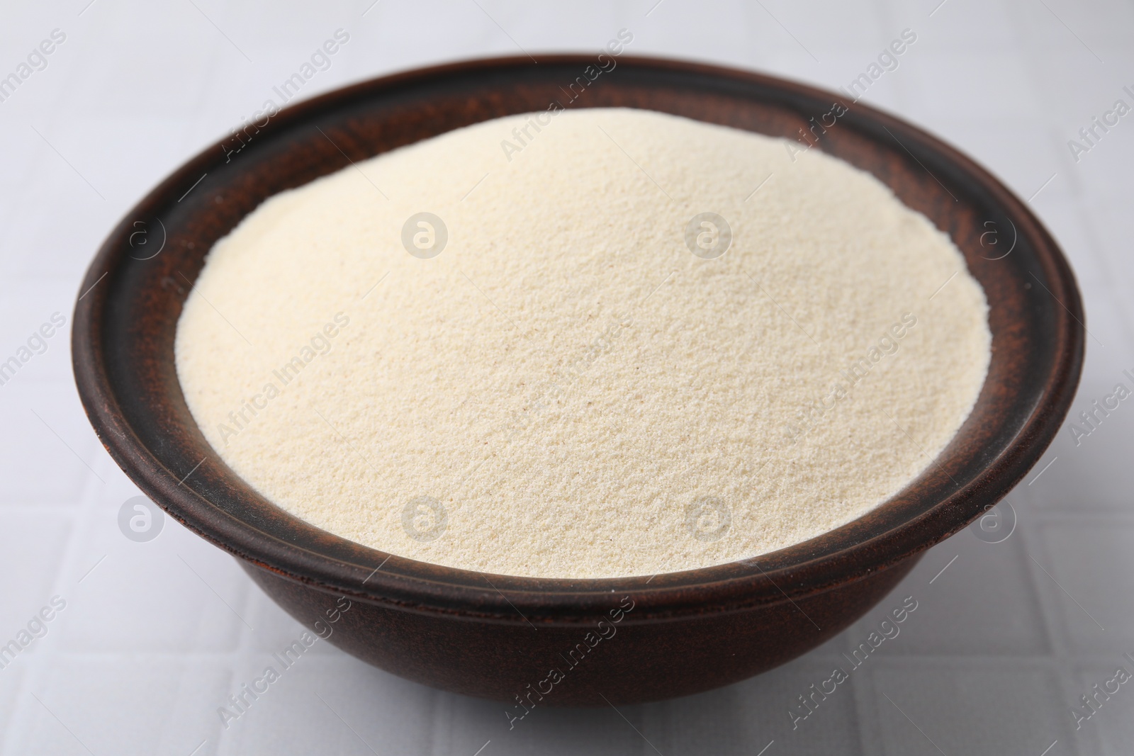 Photo of Uncooked organic semolina in bowl on white tiled table, closeup