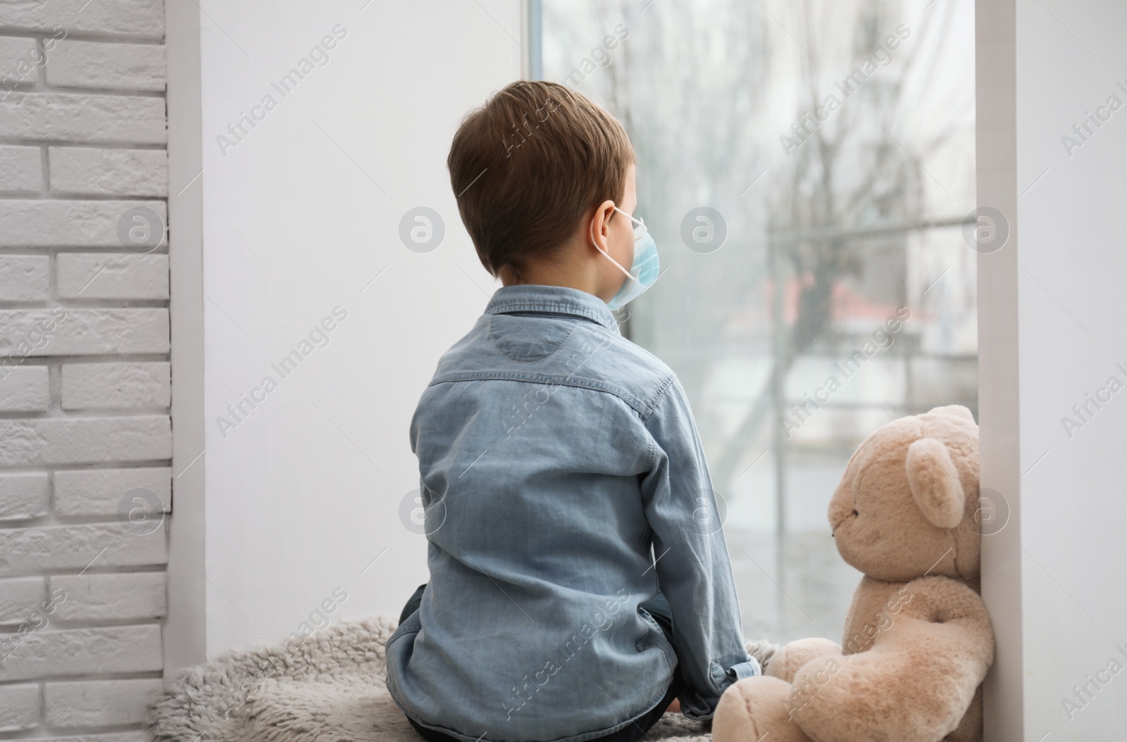 Photo of Sad little boy in protective mask looking out of window indoors. Staying at home during coronavirus pandemic