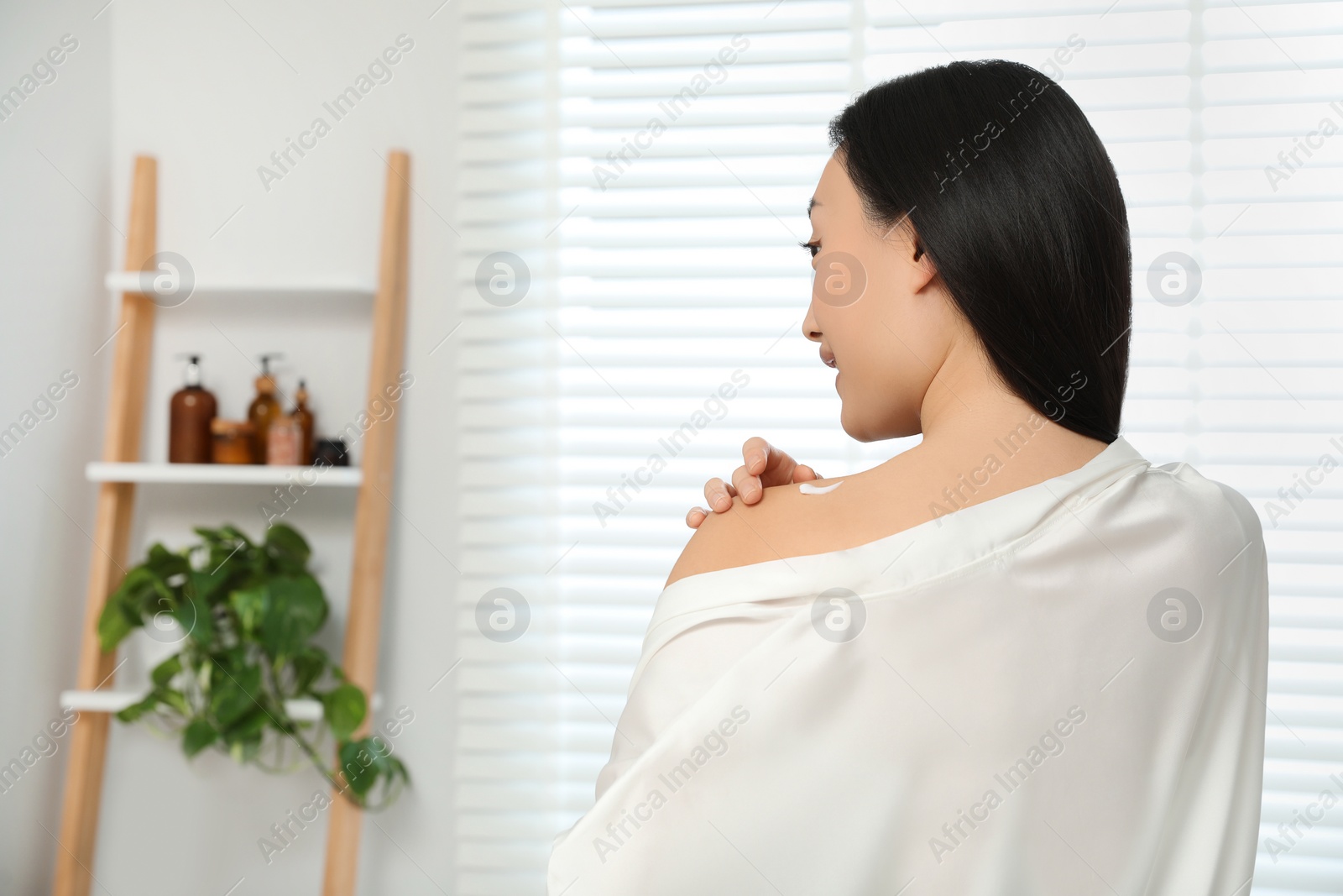 Photo of Beautiful young Asian woman applying body cream on shoulder in room, space for text