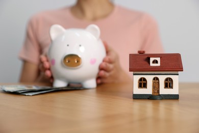 Woman holding piggy bank with banknotes at wooden table, focus on little house model