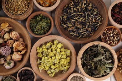 Many different dry herbs and flowers in bowls on white wooden table, flat lay