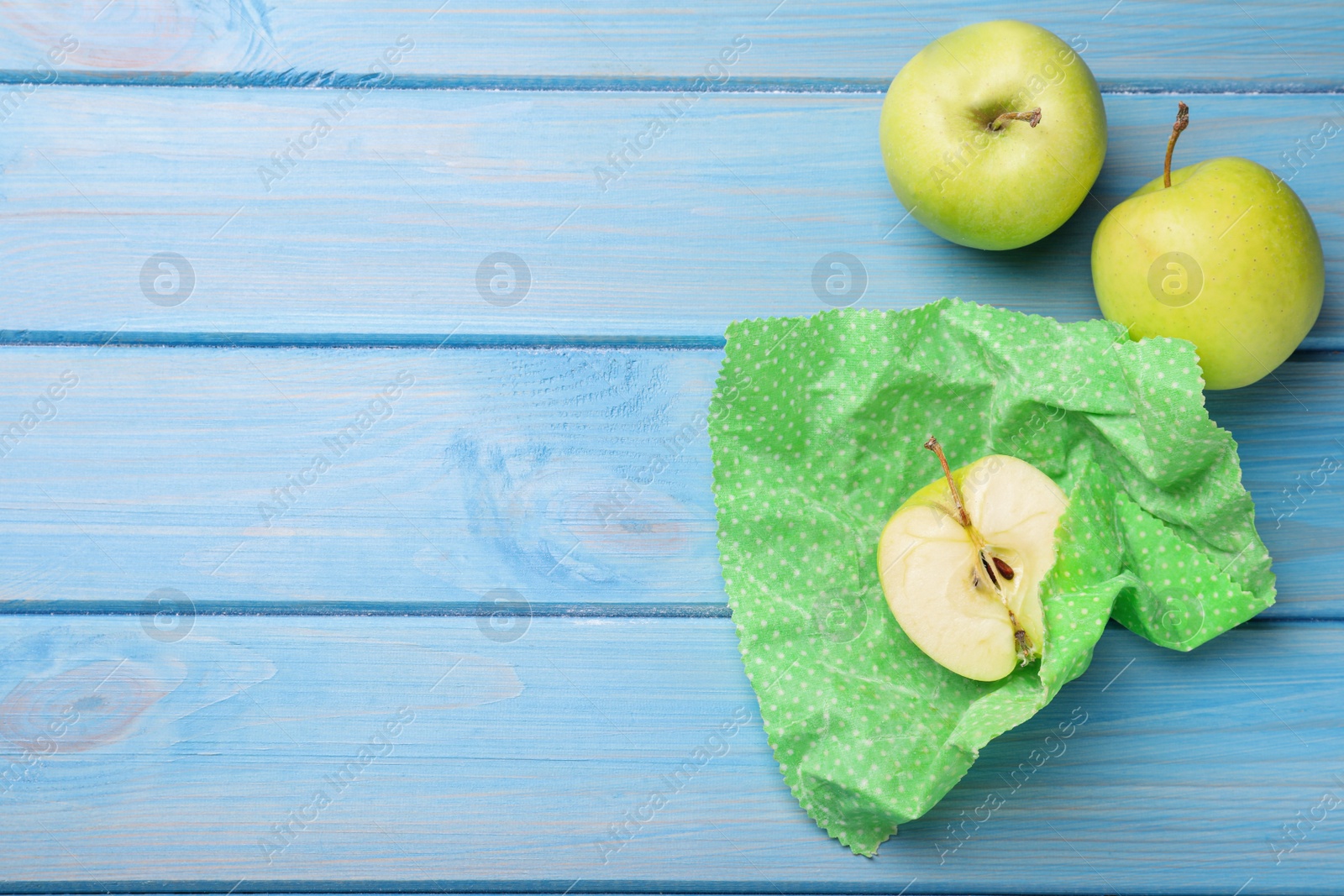 Photo of Apples with beeswax food wrap on light blue wooden table, flat lay. Space for text