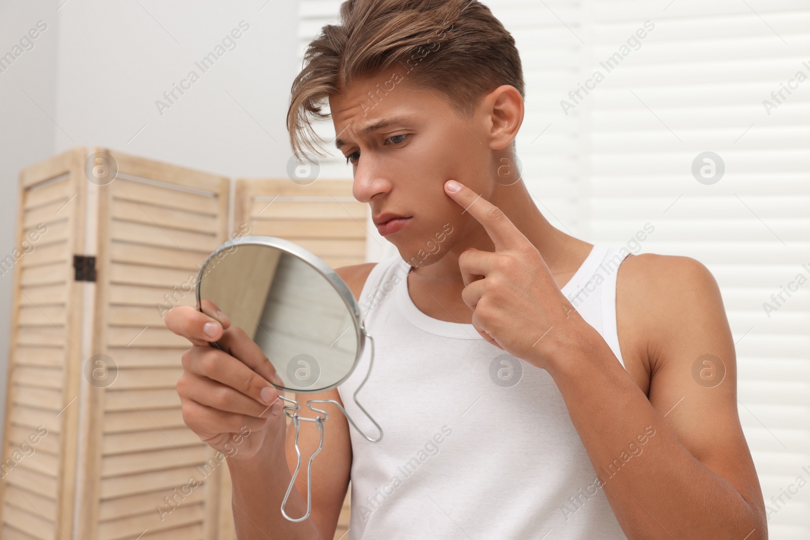 Photo of Upset young man looking at mirror and touching pimple on his face indoors. Acne problem