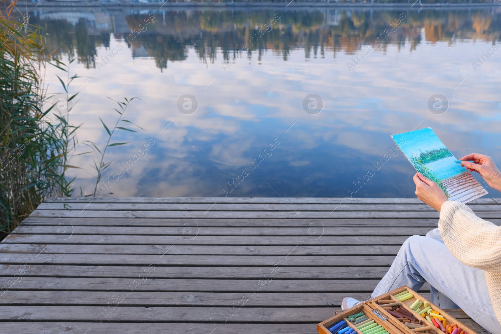 Photo of Woman drawing with soft pastels on wooden pier near river, closeup