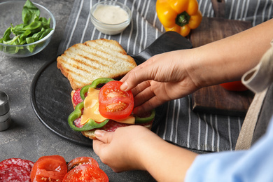 Photo of Woman adding tomato to sandwich at grey table, closeup